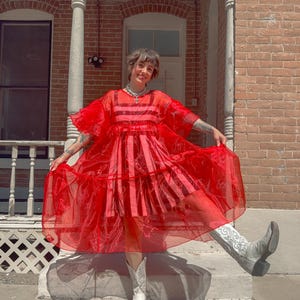 May include: A vibrant red sheer organza dress featuring short ruffled sleeves, a tiered skirt, and a square neckline. The dress is worn over a matching red and pink striped sleeveless undergarment. The outfit is paired with white cowboy boots.