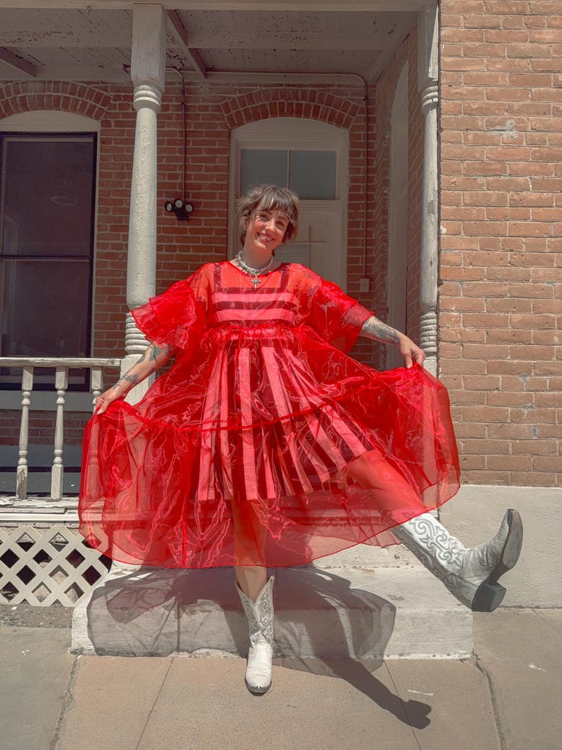 May include: A vibrant red sheer organza dress featuring short ruffled sleeves, a tiered skirt, and a square neckline. The dress is worn over a matching red and pink striped sleeveless undergarment. The outfit is paired with white cowboy boots.