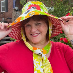 Op de afbeelding: Een kleurrijke zonnehoed met brede rand, versierd met een bloemenpatroon in rood, geel en groen. De onderkant van de hoed heeft een rood-wit ruitpatroon. Een bijpassende sjaal met bloemenmotief is geknoopt.