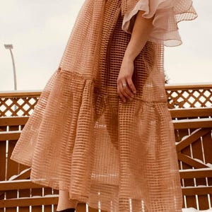 May include: A sheer, light brown midi dress featuring a grid pattern, tiered skirt, and layered ruffled sleeves. The dress is worn with dark brown ankle boots with block heels. The garment has a loose, flowing silhouette and a semi-transparent texture.