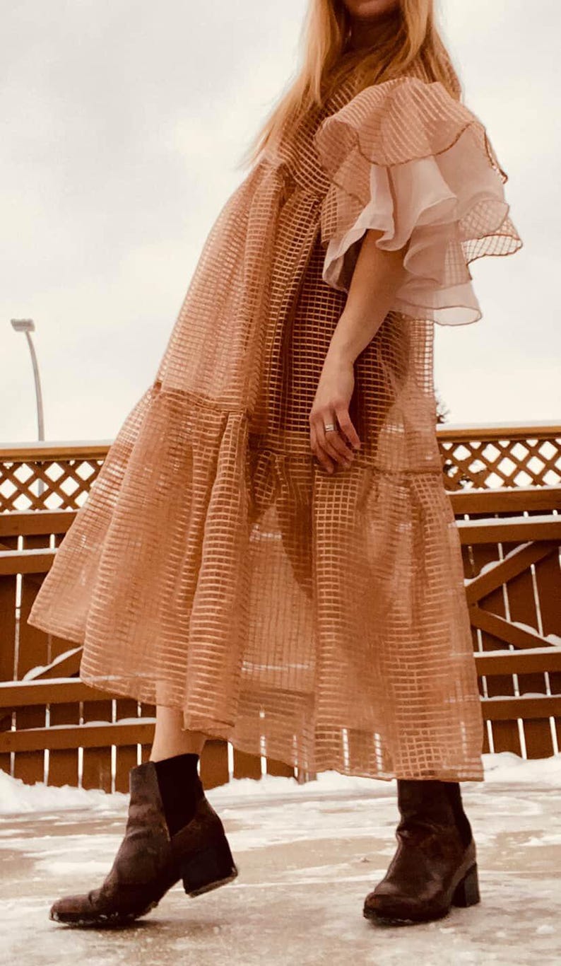 May include: A sheer, light brown midi dress featuring a grid pattern, tiered skirt, and layered ruffled sleeves. The dress is worn with dark brown ankle boots with block heels. The garment has a loose, flowing silhouette and a semi-transparent texture.