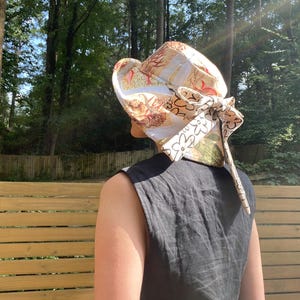 Op de afbeelding: Een zonnehoed met patroon met een brede rand en een decoratieve strik. De hoed heeft een mix van beige, witte en groene bloemen- en nautische patronen. De hoed wordt gedragen door een persoon die een zwarte mouwloze top draagt.