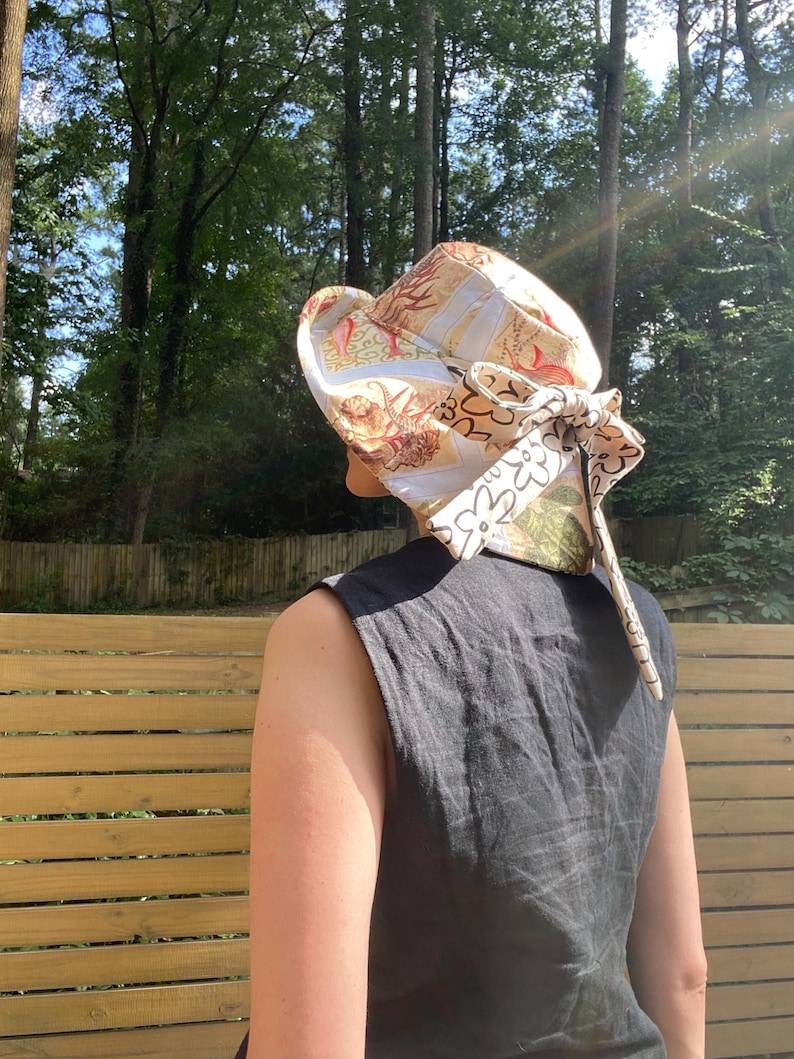 Op de afbeelding: Een zonnehoed met patroon met een brede rand en een decoratieve strik. De hoed heeft een mix van beige, witte en groene bloemen- en nautische patronen. De hoed wordt gedragen door een persoon die een zwarte mouwloze top draagt.
