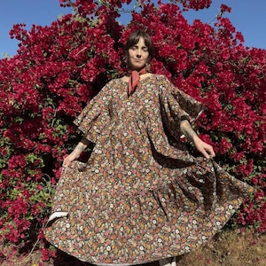 May include: A floor-length dress featuring a dark brown base with a dense, multi-colored floral pattern, wide flutter sleeves, a gathered waistline, and a tiered skirt, paired with a small orange neck scarf and white leather cowboy boots.