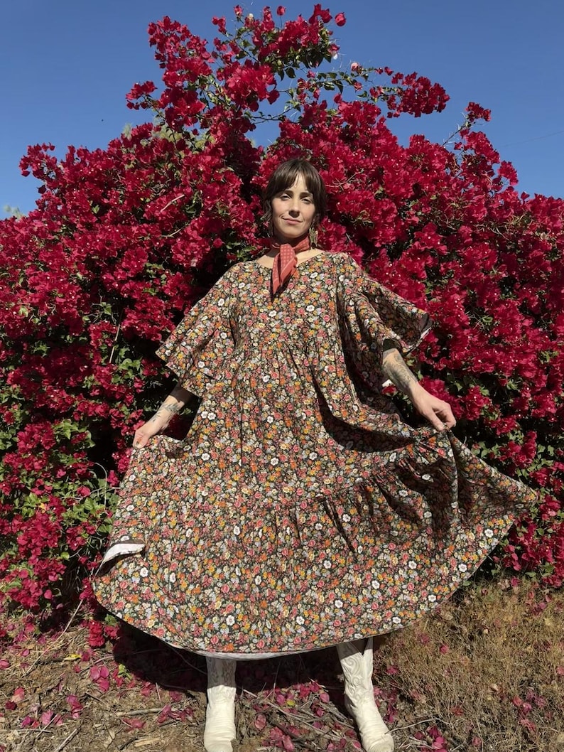 May include: A floor-length dress featuring a dark brown base with a dense, multi-colored floral pattern, wide flutter sleeves, a gathered waistline, and a tiered skirt, paired with a small orange neck scarf and white leather cowboy boots.