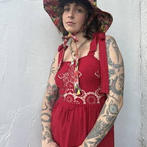 Op de afbeelding: Rode jurk met een bloemen kanten lijfje en een bijpassende breedgerande hoed. De hoed en jurk hebben een bloemenpatroon met roze, gele en rode bloemen. De jurk heeft strikbandjes en een haakontwerp.