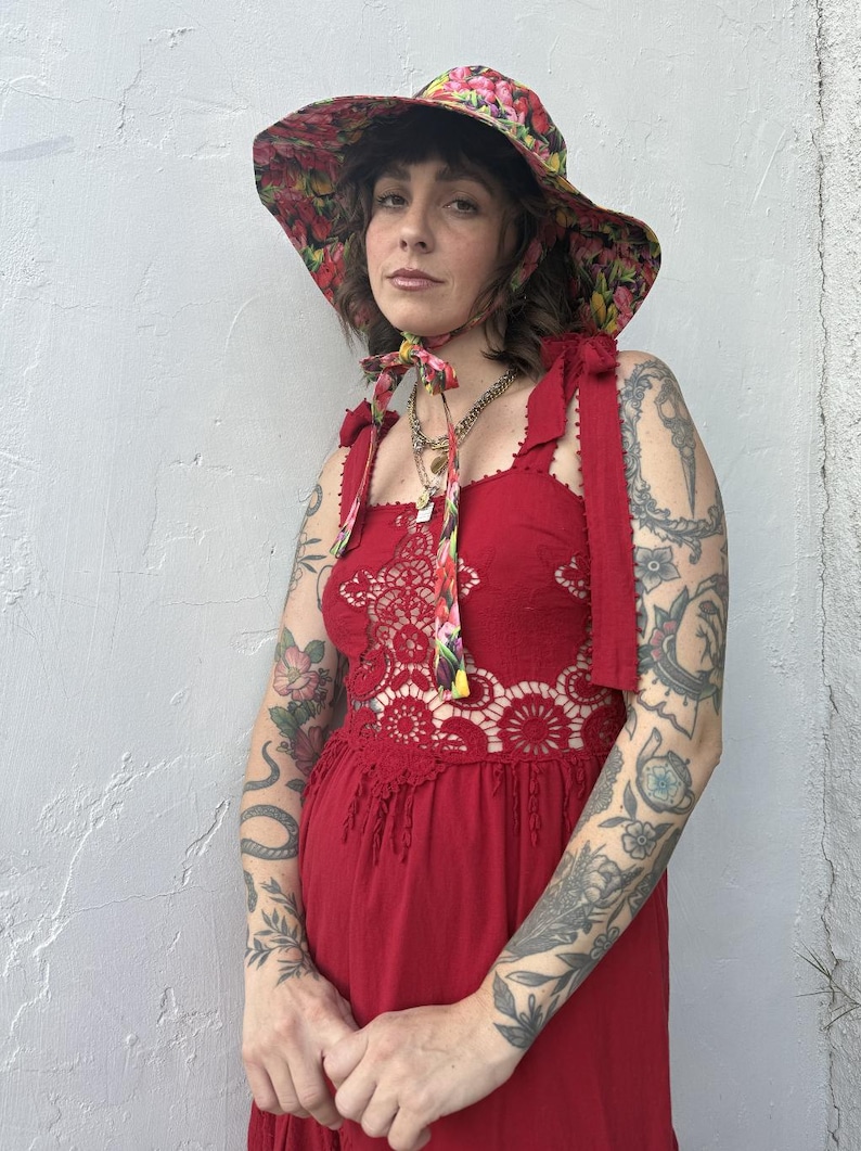 Op de afbeelding: Rode jurk met een bloemen kanten lijfje en een bijpassende breedgerande hoed. De hoed en jurk hebben een bloemenpatroon met roze, gele en rode bloemen. De jurk heeft strikbandjes en een haakontwerp.