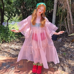May include: A sheer, light pink, tiered midi dress with voluminous ruffled sleeves, worn over a dark green top and red bottoms. The outfit is paired with red, strawberry-patterned platform shoes featuring green leaf-shaped cuffs at the ankles.