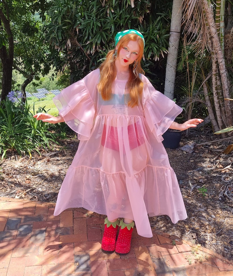 May include: A sheer, light pink, tiered midi dress with voluminous ruffled sleeves, worn over a dark green top and red bottoms. The outfit is paired with red, strawberry-patterned platform shoes featuring green leaf-shaped cuffs at the ankles.