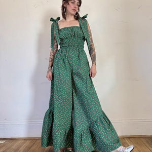 May include: A green jumpsuit with a floral pattern. The jumpsuit features a square neckline, tie straps, and a shirred bodice. The wide-leg pants have a ruffled hem. The model is wearing silver shoes.