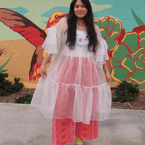 May include: A white sheer tiered dress with ruffled sleeves worn over a white graphic tank top and wide-leg trousers featuring a pink and orange geometric pattern. The outfit is completed with yellow socks and metallic silver block-heeled shoes.