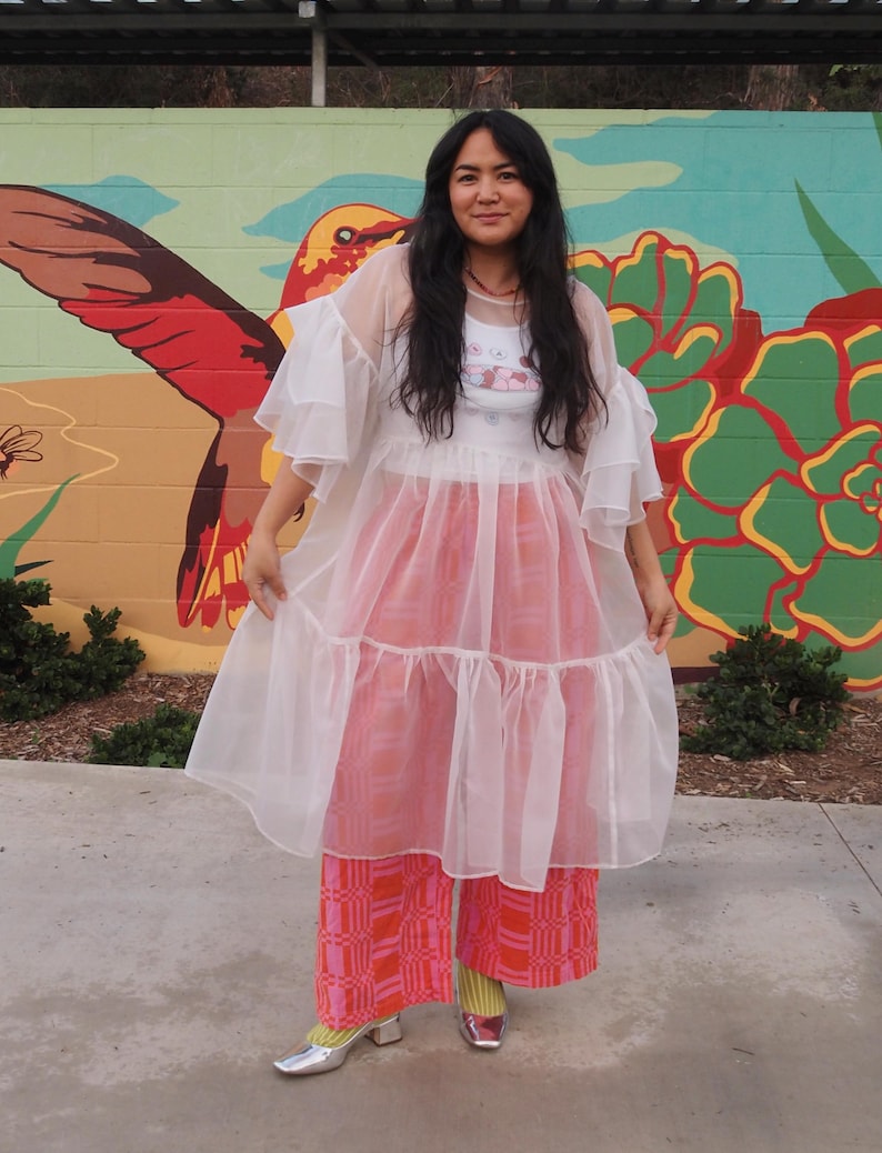 May include: A white sheer tiered dress with ruffled sleeves worn over a white graphic tank top and wide-leg trousers featuring a pink and orange geometric pattern. The outfit is completed with yellow socks and metallic silver block-heeled shoes.