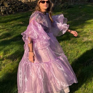 May include: A sheer lavender organza dress featuring voluminous tiered ruffled sleeves and a loose, flowing silhouette. The garment is worn over light-colored trousers and dark shoes, showcasing a translucent, lightweight fabric with a subtle sheen.