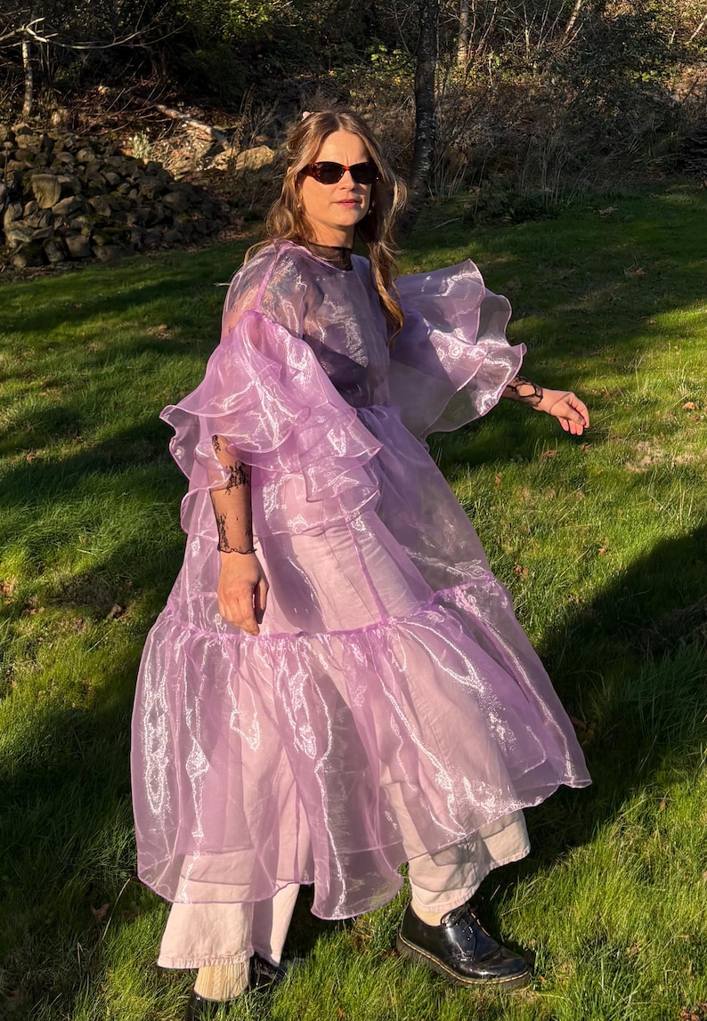 May include: A sheer lavender organza dress featuring voluminous tiered ruffled sleeves and a loose, flowing silhouette. The garment is worn over light-colored trousers and dark shoes, showcasing a translucent, lightweight fabric with a subtle sheen.