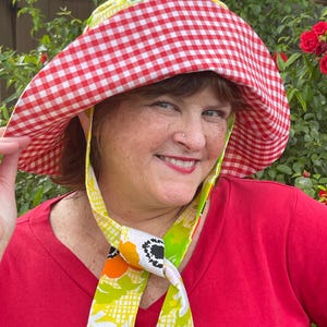 Op de afbeelding: Een zonnehoed met een rood-wit ruitpatroon en een brede rand. De hoed heeft een bloemenpatroon en wordt gedragen met een rode top. De hoed is ontworpen voor bescherming tegen de zon.