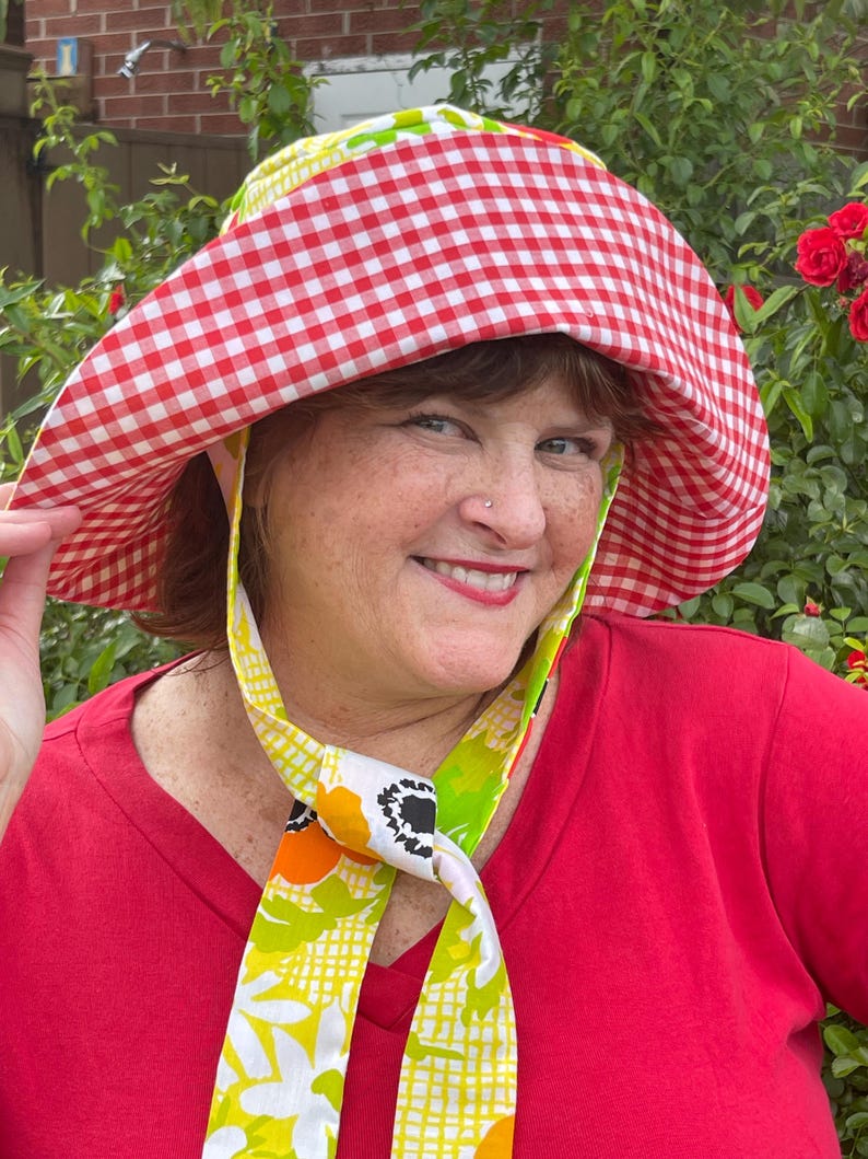 Op de afbeelding: Een zonnehoed met een rood-wit ruitpatroon en een brede rand. De hoed heeft een bloemenpatroon en wordt gedragen met een rode top. De hoed is ontworpen voor bescherming tegen de zon.