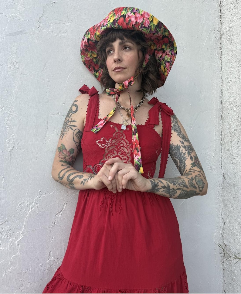 Op de afbeelding: Rode zomerjurk met een bloemen kanten lijfje en een rok met ruches. De jurk heeft strikbandjes en wordt gecombineerd met een breedgerande bloemenhoed met een bijpassende stropdas. De hoed heeft een levendige mix van roze, gele en rode bloemen.