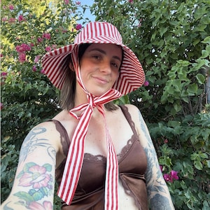 Op de afbeelding: Een zonnehoed met brede rand in rood en wit gestreept, met bijpassende linten. De hoed wordt gedragen met een bruine satijnen top en een rok met luipaardprint. De achtergrond toont groene bladeren en roze bloemen.