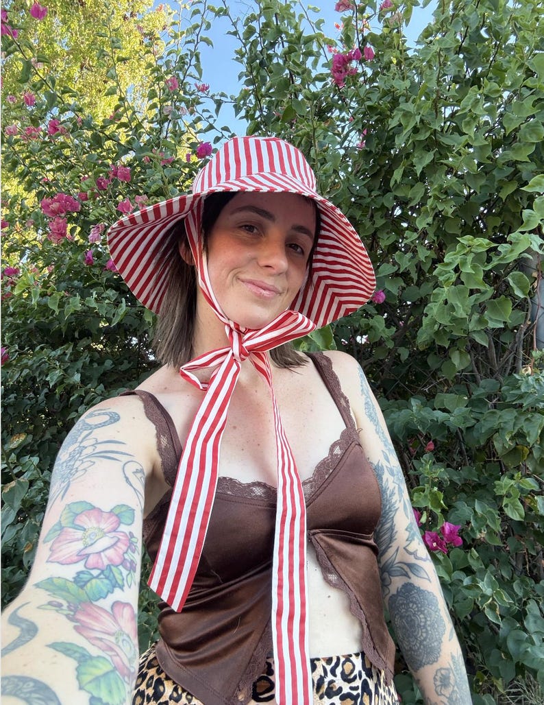 Op de afbeelding: Een zonnehoed met brede rand in rood en wit gestreept, met bijpassende linten. De hoed wordt gedragen met een bruine satijnen top en een rok met luipaardprint. De achtergrond toont groene bladeren en roze bloemen.