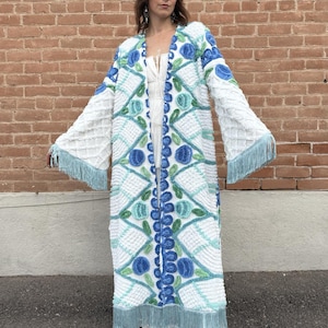 May include: A long, white, textured duster coat featuring a blue and green floral pattern with diamond-shaped stitching. The garment has wide, long sleeves and a light blue fringe trim along the hemline and the edges of the sleeve cuffs.
