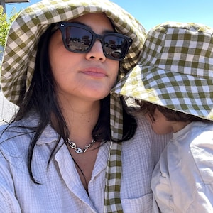 Op de afbeelding: Een vrouw en kind dragen bijpassende groen-witte geruite vissershoeden en een zwarte zonnebril. De vrouw draagt ook een zilveren ketting en een wit overhemd met knopen. Het kind draagt een wit shirt.
