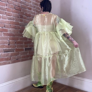 May include: A sheer, pale lime green organza duster robe featuring voluminous, multi-layered ruffled sleeves and a tiered skirt design. The garment is worn over a matching light yellow camisole and shorts set, paired with green floral cowboy boots.