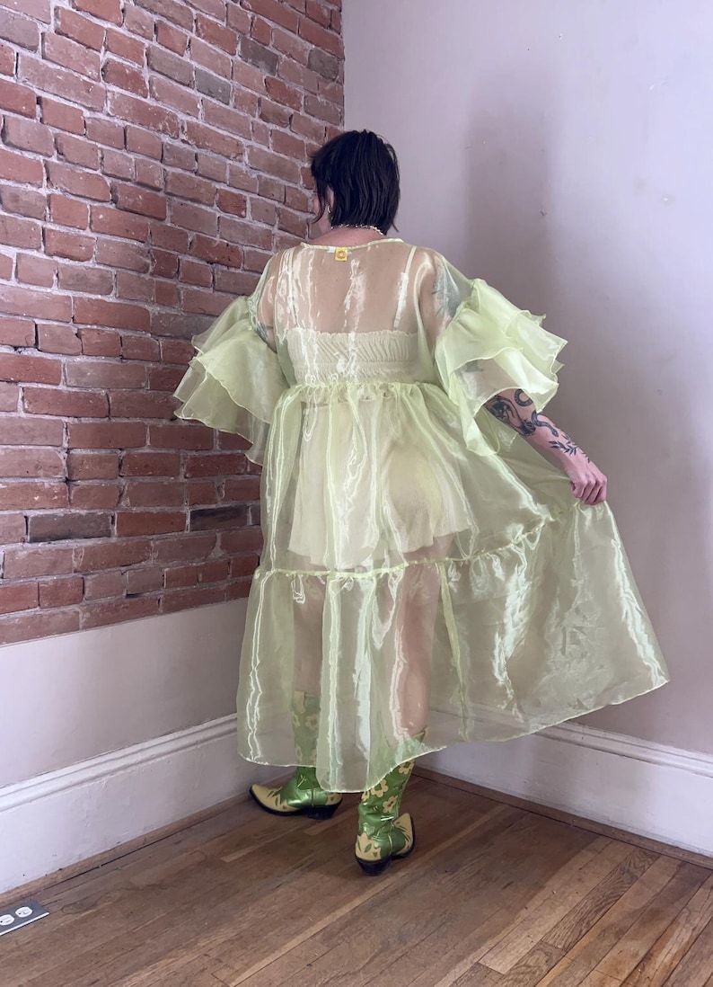 May include: A sheer, pale lime green organza duster robe featuring voluminous, multi-layered ruffled sleeves and a tiered skirt design. The garment is worn over a matching light yellow camisole and shorts set, paired with green floral cowboy boots.