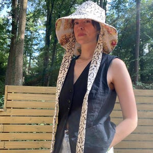 Op de afbeelding: Een zonnehoed met brede rand met een witte achtergrond en een herhalend zwart bloemenpatroon. De hoed heeft lange, bedrukte banden en een bedrukte onderkant. De persoon draagt een zwart vest en een lichtblauwe spijkerbroek.