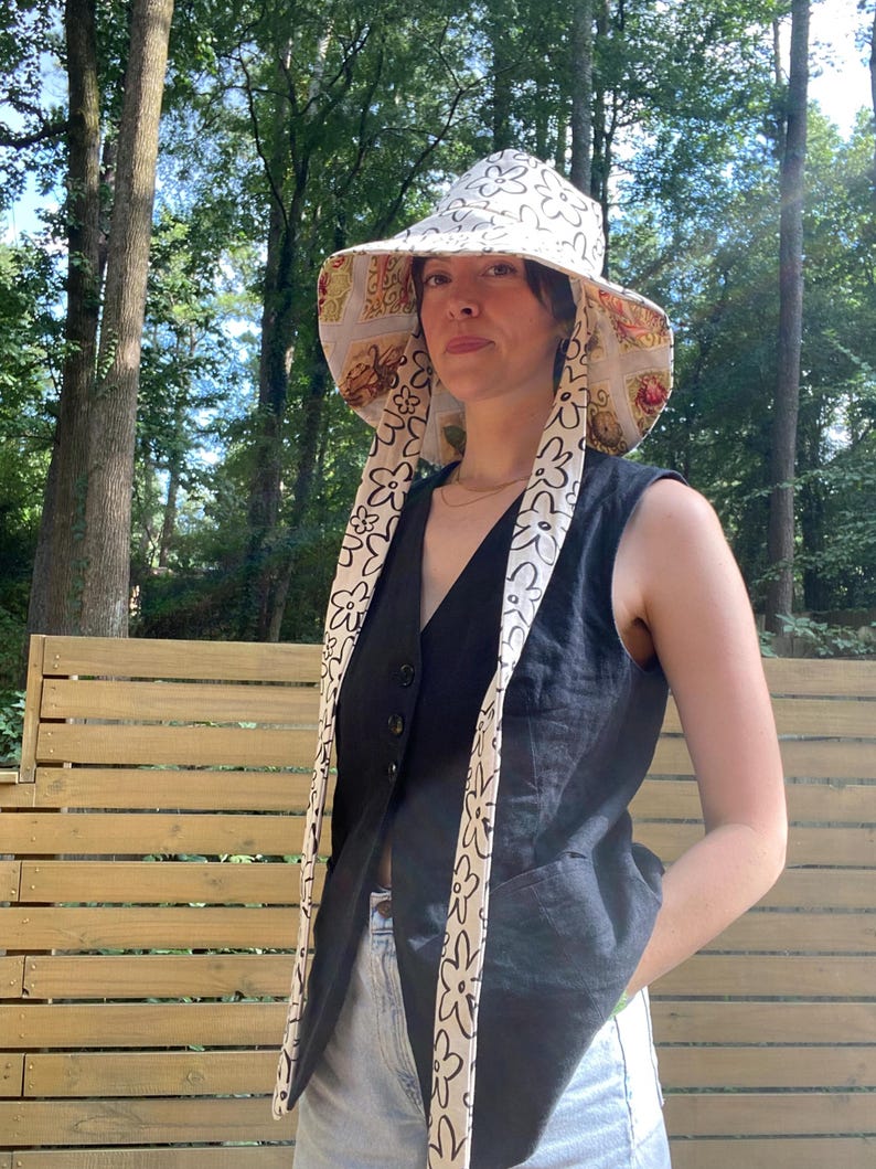 Op de afbeelding: Een zonnehoed met brede rand met een witte achtergrond en een herhalend zwart bloemenpatroon. De hoed heeft lange, bedrukte banden en een bedrukte onderkant. De persoon draagt een zwart vest en een lichtblauwe spijkerbroek.