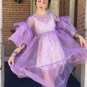 May include: A sheer, lavender-colored organza dress featuring voluminous, ruffled sleeves and a tiered skirt. The dress is worn over a light pink, sleeveless lace slip dress. The garment has a round neckline and a loose, flowing silhouette.