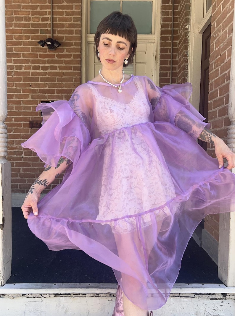 May include: A sheer, lavender-colored organza dress featuring voluminous, ruffled sleeves and a tiered skirt. The dress is worn over a light pink, sleeveless lace slip dress. The garment has a round neckline and a loose, flowing silhouette.