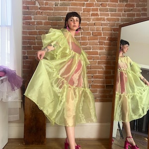 May include: A sheer, lime green organza dress featuring voluminous ruffled sleeves and a tiered skirt design. The garment is worn over a solid pink slip dress and is paired with bright pink platform sandals with bow details and matching earrings.
