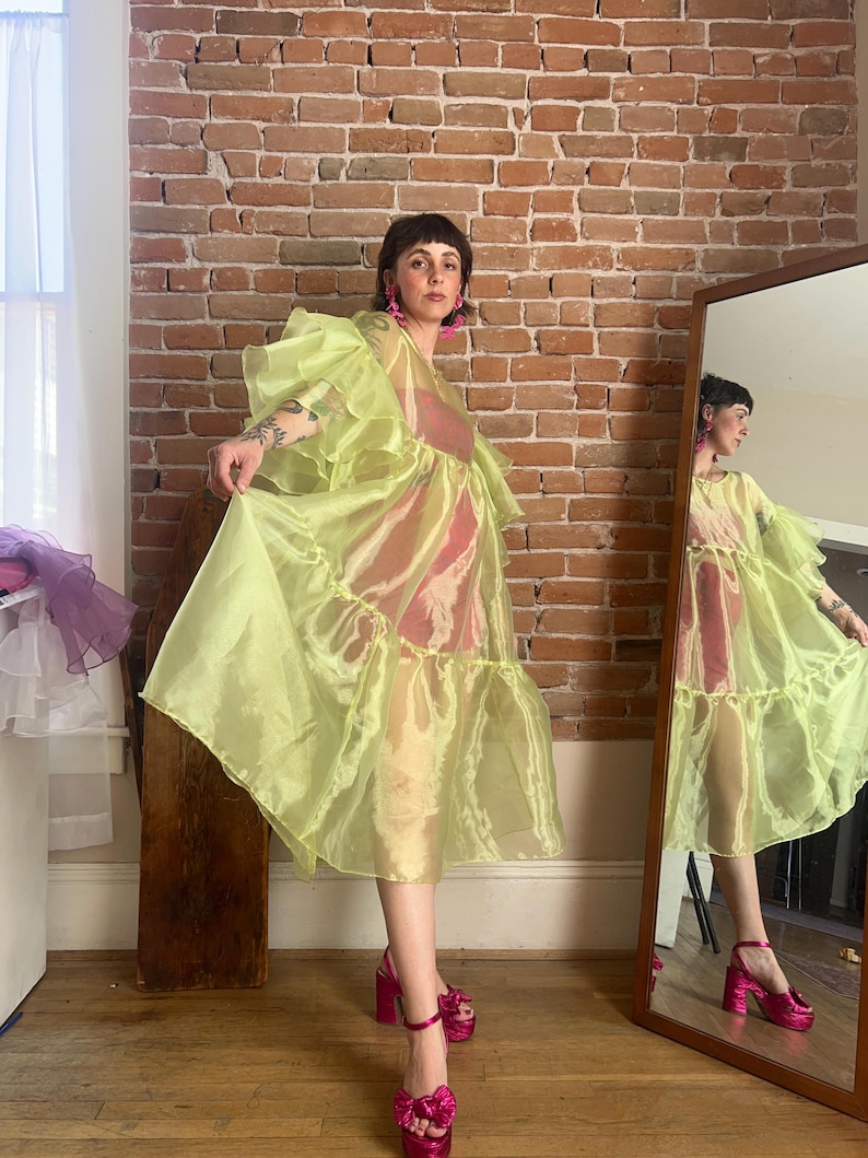 May include: A sheer, lime green organza dress featuring voluminous ruffled sleeves and a tiered skirt design. The garment is worn over a solid pink slip dress and is paired with bright pink platform sandals with bow details and matching earrings.