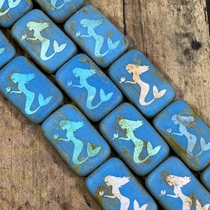 May include: Rectangular blue beads with a mermaid design. The mermaids are in various colors, including white, light blue, and peach. The beads have a distressed, vintage look, and are arranged diagonally.