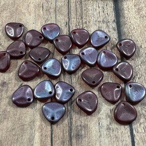 May include: A collection of maroon-colored, triangular beads with iridescent blue and purple highlights. Each bead has a hole for stringing and a textured surface. The beads are scattered on a wooden surface.