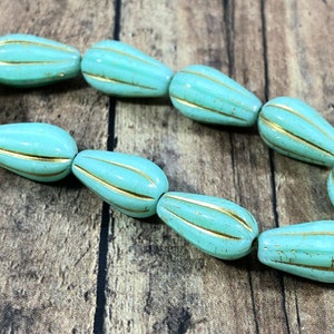 May include: A strand of turquoise teardrop-shaped beads with gold stripes. The beads are strung together, showcasing a repeating pattern. The beads have a glossy finish and are arranged on a wooden surface.