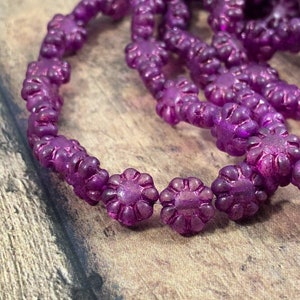May include: A strand of purple glass beads with a flower design. The beads are iridescent and have a frosted finish.