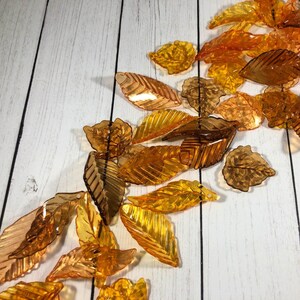 May include: A collection of translucent amber leaf-shaped ornaments, scattered on a white wood background.