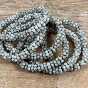 May include: Several coiled bracelets made of small, round, light gray beads with silver-colored accents. The bracelets are arranged on a light brown wooden surface, creating a textured and layered effect.