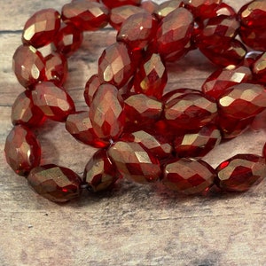 May include: Strand of faceted, oval-shaped red beads. The beads have a glossy finish and are strung together, creating a curved shape. The beads are resting on a wooden surface.
