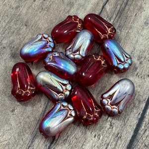 May include: A collection of artisan beads in shades of red and iridescent blue. The beads have a tulip-like shape with detailed petal designs and a glossy finish. The beads are scattered on a gray surface.