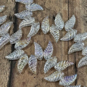 May include: A collection of clear, iridescent leaf-shaped beads. Each bead has a detailed vein pattern and reflects light with a rainbow sheen. The beads are scattered on a wooden surface, creating a decorative element.