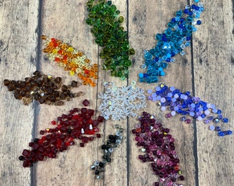 May include: A collection of colorful faceted beads arranged in a starburst pattern. The beads are in shades of red, brown, orange, green, blue, purple, clear, and black. The beads are on a wooden surface.
