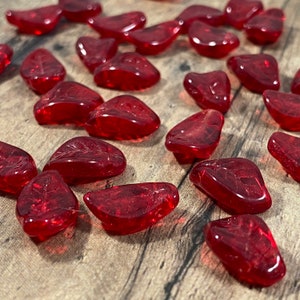 May include: A collection of translucent, heart-shaped, red glass stones scattered on a wooden surface. The stones vary slightly in size and shape, with smooth, rounded edges. The lighting creates highlights and shadows, enhancing their glossy appearance.