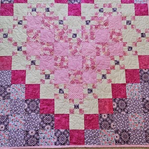 May include: A handmade quilt featuring a heart design. The quilt showcases a patchwork of pink, purple, and cream squares, with a heart shape in the center. The heart is made of floral patterned fabric and quilted with a floral design.
