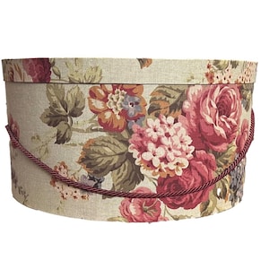 Può includere: Scatola ovale con stampa floreale, con sfondo crema e un motivo di rose rosse e rosa, foglie verdi e altri fiori. La scatola ha un manico in corda bordeaux.