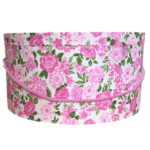 Könnte beinhalten: Eine runde, zylindrische Hutschachtel mit weißem Hintergrund und einem dichten Muster aus rosa Rosen und grünen Blättern. Die Schachtel verfügt über einen passenden, gedrehten rosa Kordelgriff an den Seiten, der als Tragehilfe dient.