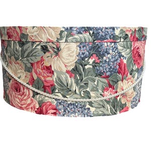 Puede incluir: Una caja de almacenamiento redonda con estampado floral y un asa de cuerda blanca. La caja presenta un patrón repetido de rosas rosas, hortensias azules y hojas verdes sobre un fondo crema.