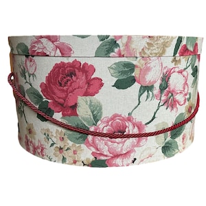 Può includere: Una cappelliera rotonda floreale con coperchio. La scatola è rivestita in tessuto color crema con un motivo di rose rosa e rosse, foglie verdi e piccoli fiori gialli. È presente un manico in corda rossa.