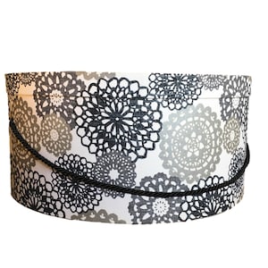 May include: A round hat box with a black rope handle. The box is decorated with a white fabric featuring a repeating pattern of grey and black floral designs. The box is approximately 25 cm in diameter.